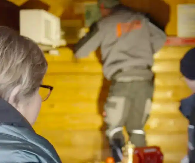 Person in Winterkleidung betreut Szene an Holzstall, während andere Person mit Werkzeug hantiert.