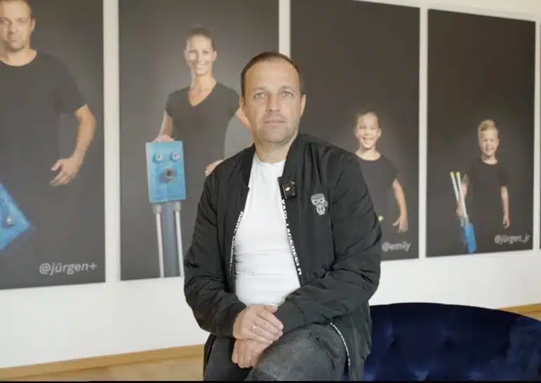 Jürgen Höfler sitzt auf einem Sofa vor großen Porträts in einem hellen Raum, möglicherweise Imagevideo-Set.