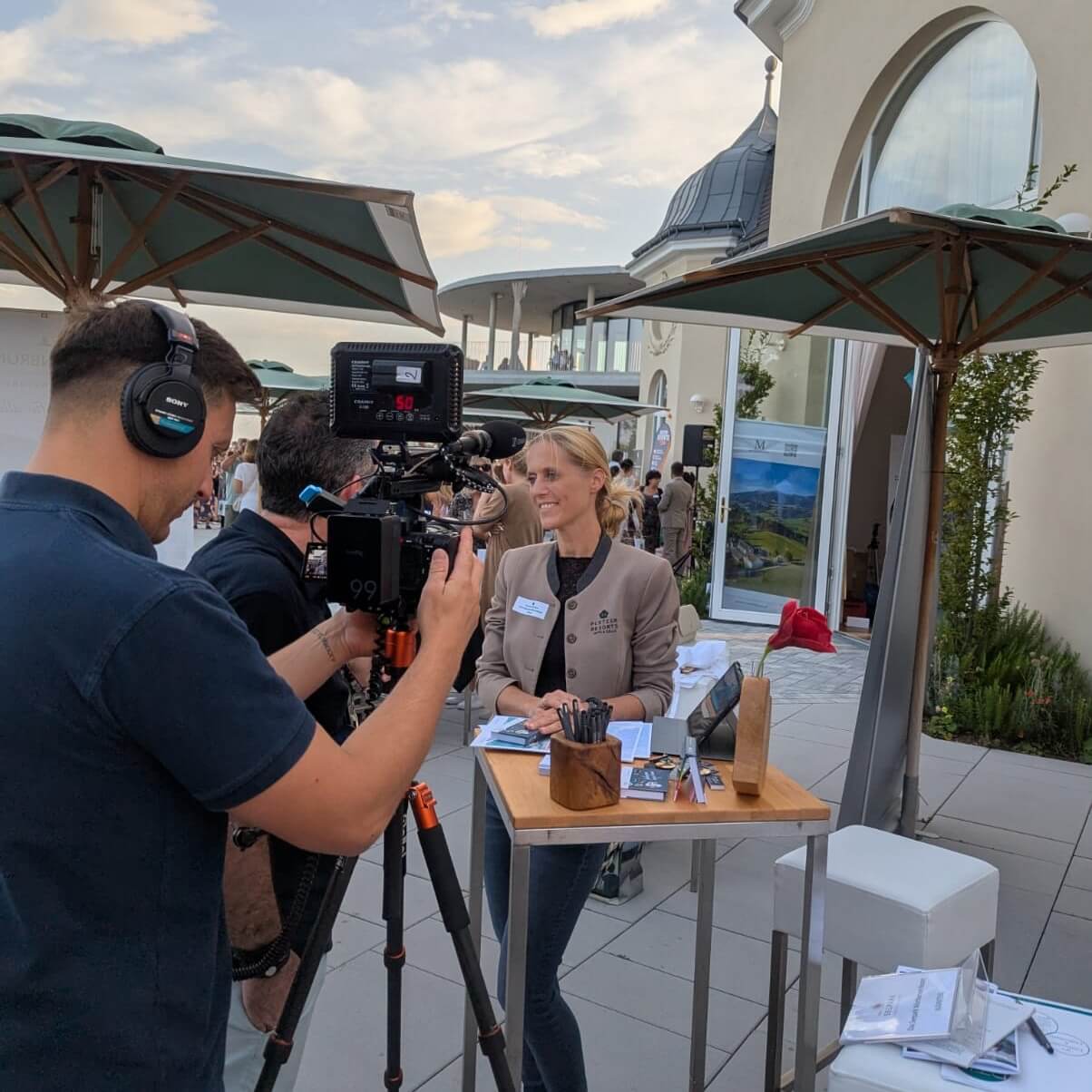 Person wird bei einem Interview im Freien gefilmt, vor einem Stand mit Broschüren, unter Sonnenschirmen bei einer Veranstaltung.
