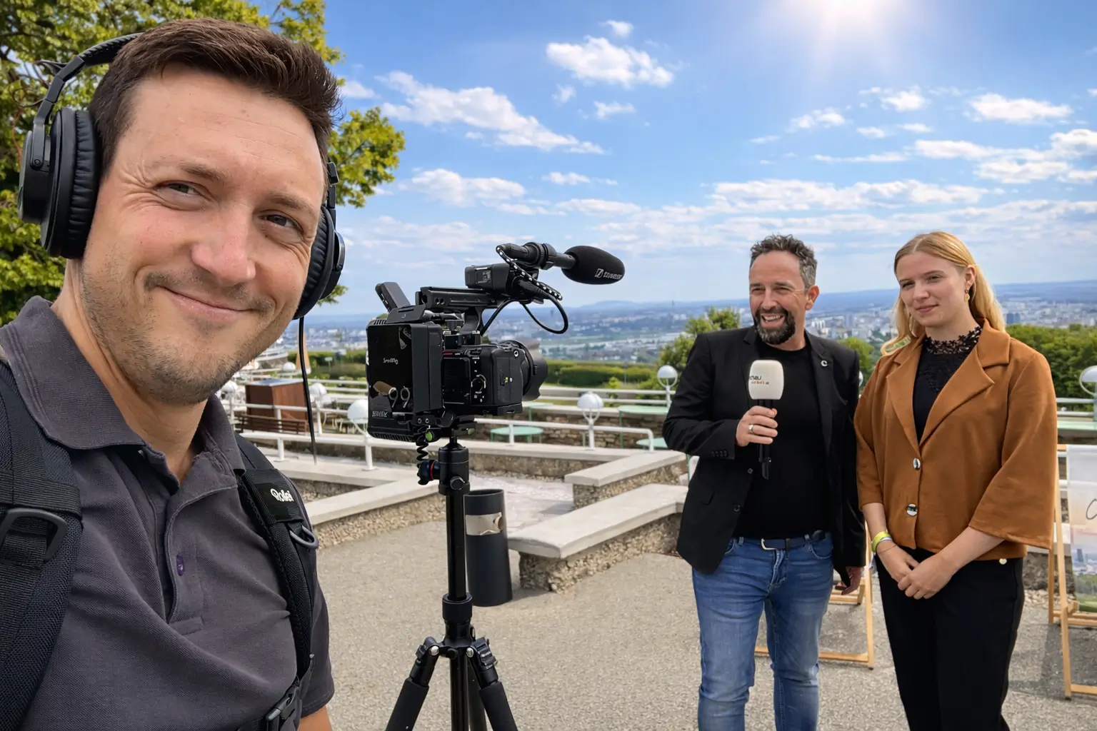 Person mit Kopfhörern und Kamera bei Interview im Freien; zwei weitere Personen stehen vor Mikrofon mit Stadtblick.