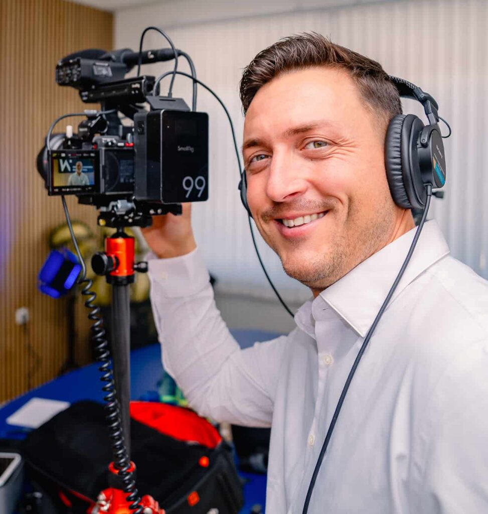 Person mit Kopfhörern bedient Kamera bei einem Filmdreh, während eine andere Person im Hintergrund posiert.